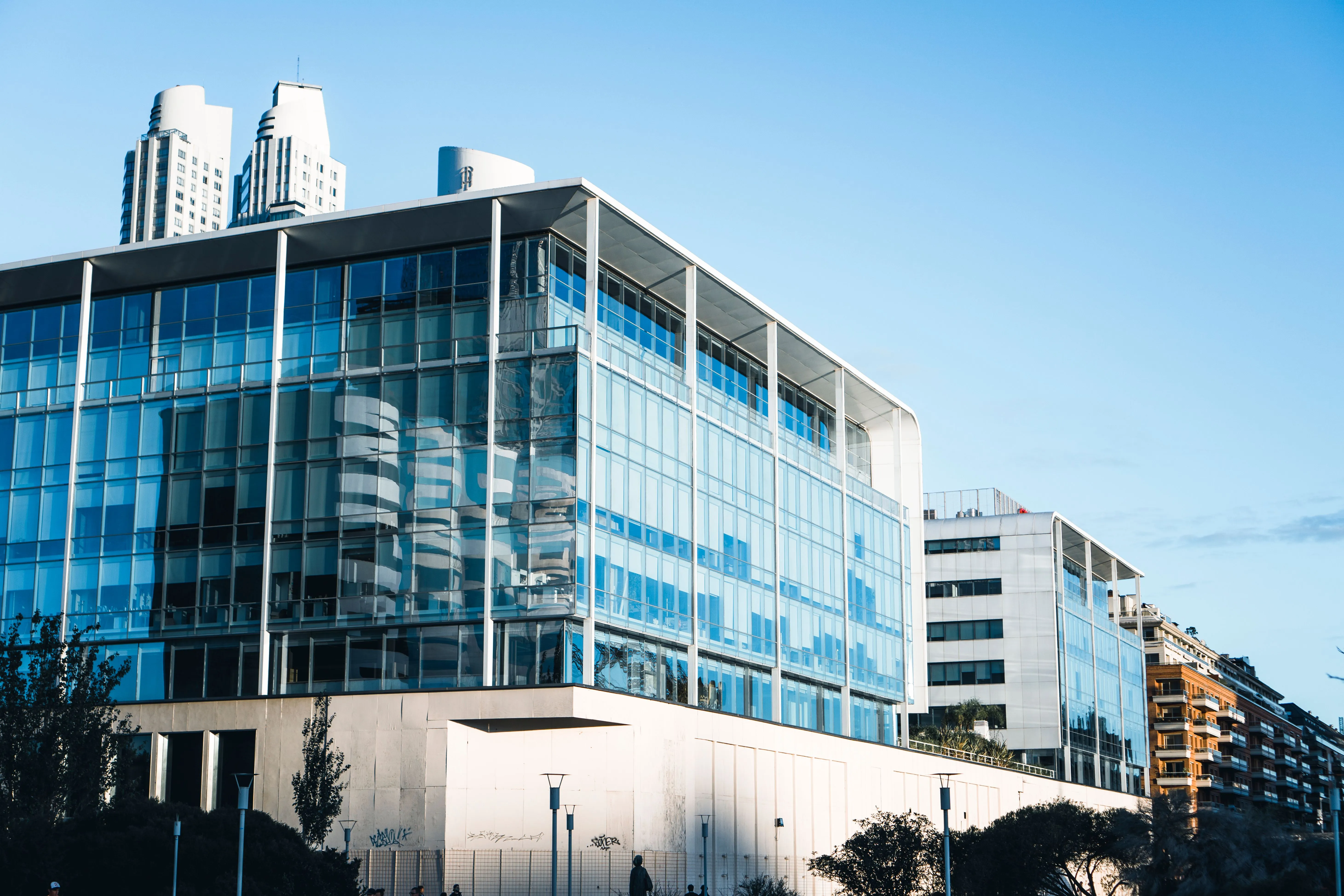 A glazed office building against clear sky, towers rising behind it.
