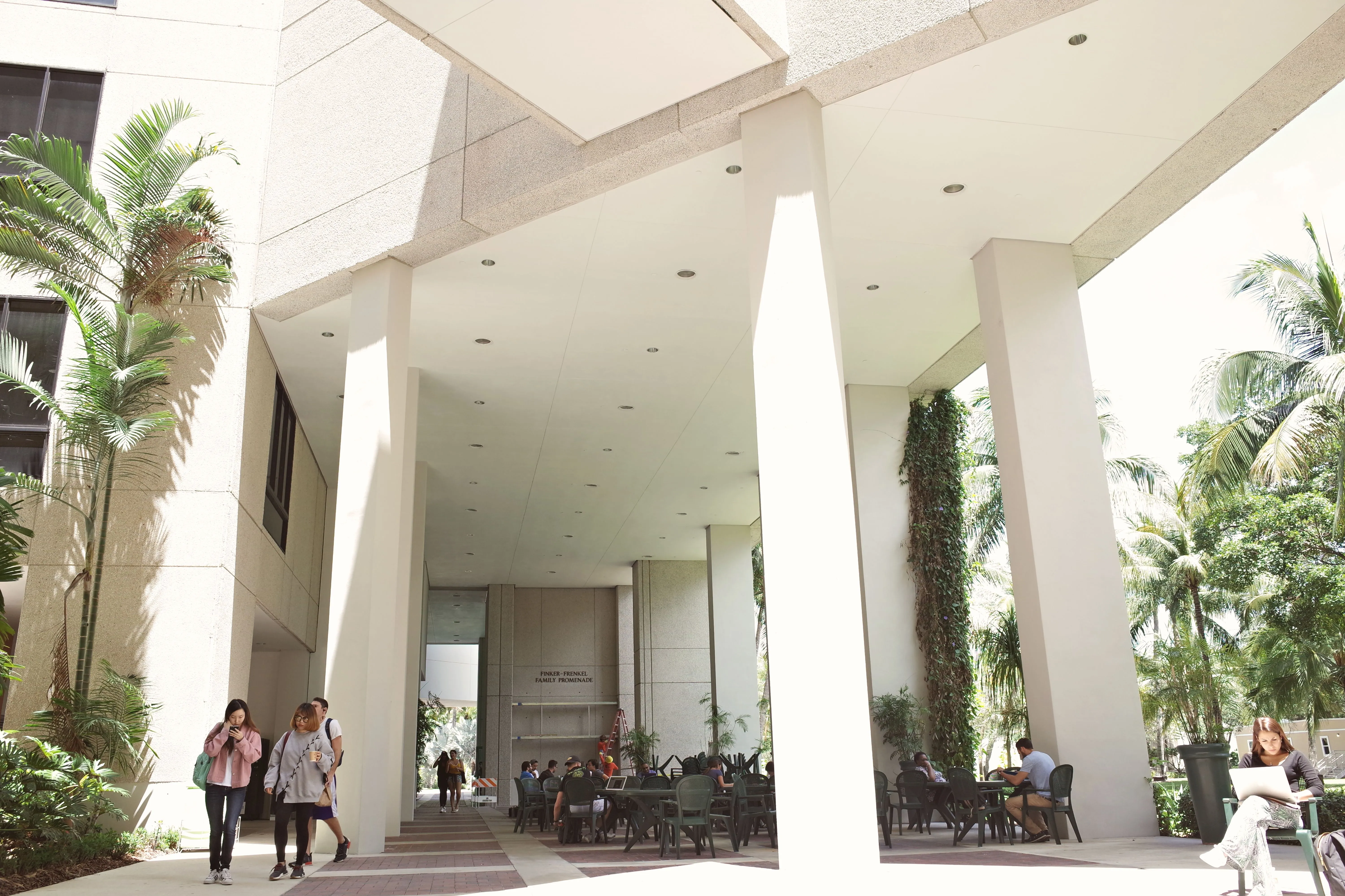 Students crossing a shaded campus walkway past outdoor study tables.